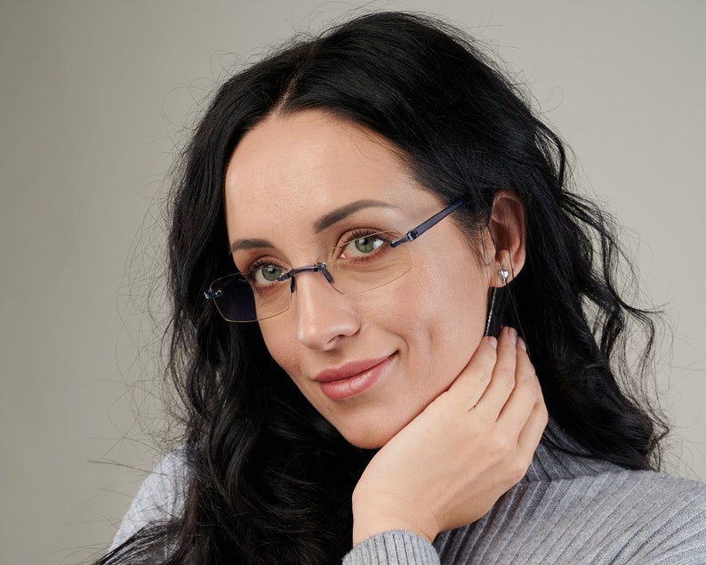Puede incluir: Una mujer con el pelo largo y negro que lleva gafas sin montura con monturas azules. Lleva un jersey gris y tiene la mano en la barbilla.