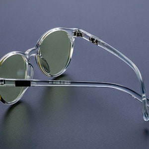 May include: Clear plastic framed round sunglasses with green tinted lenses. The sunglasses have a black accent on the bridge of the nose and the text "AERO 63 18 135" on the arm.