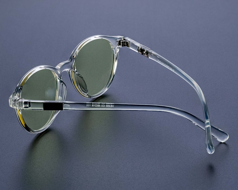 May include: Clear plastic framed round sunglasses with green tinted lenses. The sunglasses have a black accent on the bridge of the nose and the text "AERO 63 18 135" on the arm.