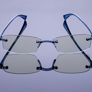 Puede incluir: Un par de gafas azules sin montura con lentes gris claro. Las gafas se reflejan en una superficie de espejo.