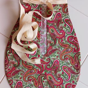 May include: A fabric bag with a green paisley pattern, featuring red, pink, and orange accents. The bag has a cream-colored ribbon closure and straps. The bag is designed with a rounded bottom and a rectangular opening.