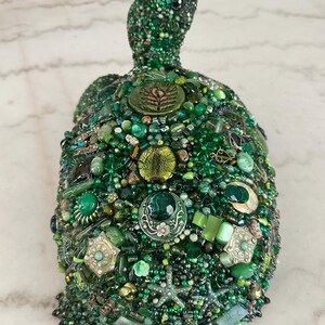 May include: A handcrafted, decorative turtle sculpture, predominantly green, adorned with a mosaic of beads, gems, and embellishments. The turtle's shell features various shades of green, creating a textured and detailed surface. The head and flippers are also covered in green beads.