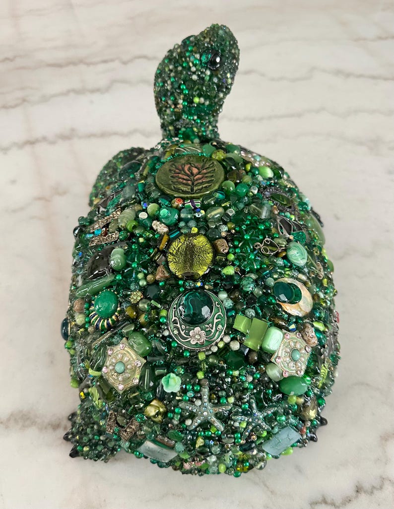 May include: A handcrafted, decorative turtle sculpture, predominantly green, adorned with a mosaic of beads, gems, and embellishments. The turtle's shell features various shades of green, creating a textured and detailed surface. The head and flippers are also covered in green beads.