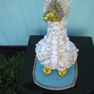 May include: A decorative goose figurine, adorned with white and iridescent embellishments. The goose has a yellow beak and feet, and blue eyes. It sits on a blue felt base with a rope border.