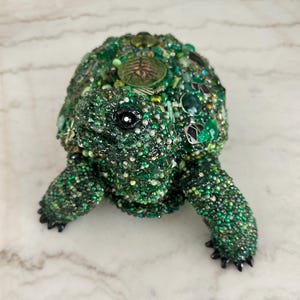 May include: A decorative turtle figurine, primarily green, adorned with beads and embellishments. The turtle's shell is covered in various shades of green beads, with accents of other colors. The figurine has a detailed head, legs, and a black eye.