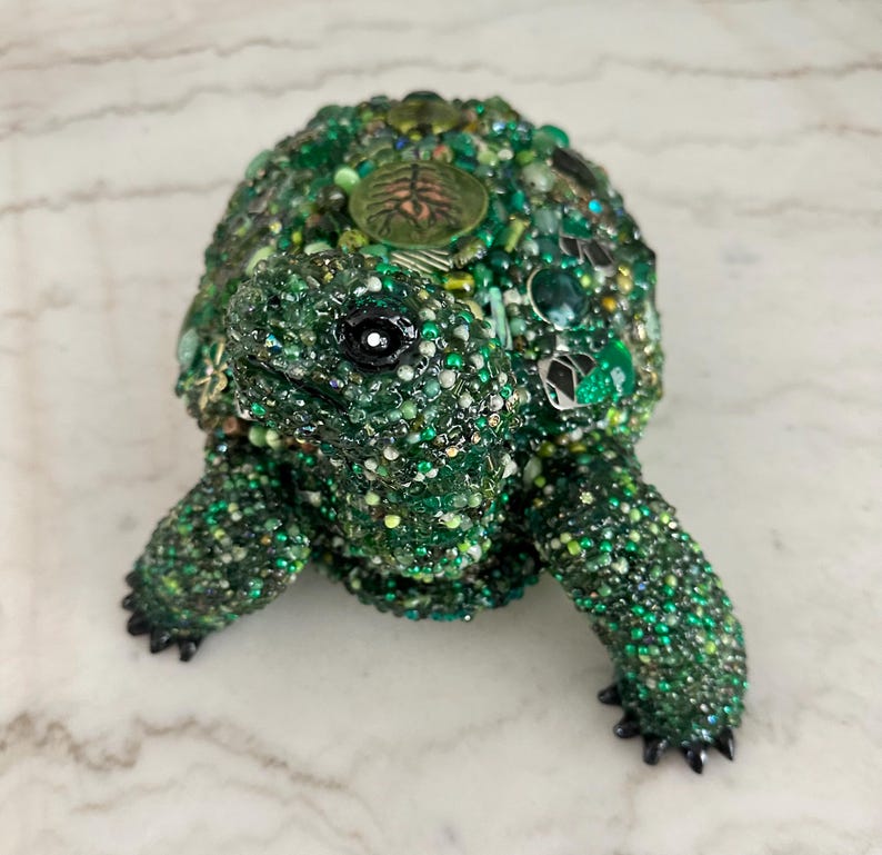 May include: A decorative turtle figurine, primarily green, adorned with beads and embellishments. The turtle's shell is covered in various shades of green beads, with accents of other colors. The figurine has a detailed head, legs, and a black eye.