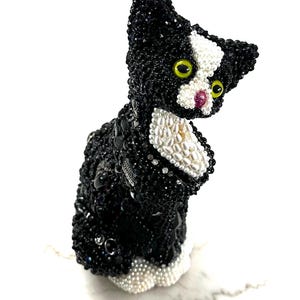 May include: A black and white beaded cat figurine with yellow eyes and a pink nose. The cat is sitting upright, with its head turned slightly to the side. The figurine is made of small, round beads and has a detailed design.
