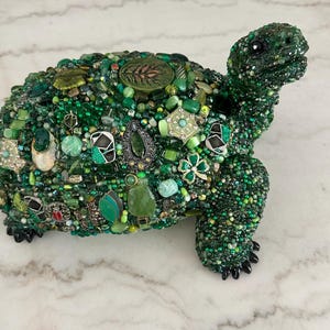 May include: A decorative turtle sculpture covered in various shades of green beads, gems, and embellishments. The turtle's shell is adorned with a variety of green elements, including beads, stones, and decorative pieces. The sculpture has black feet and a detailed head.