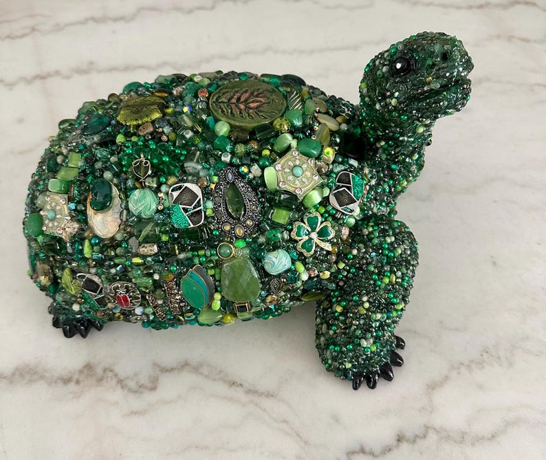 May include: A decorative turtle sculpture covered in various shades of green beads, gems, and embellishments. The turtle's shell is adorned with a variety of green elements, including beads, stones, and decorative pieces. The sculpture has black feet and a detailed head.