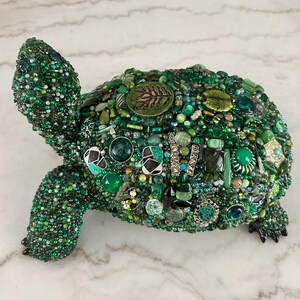 May include: A decorative turtle sculpture covered in various shades of green beads, gems, and decorative elements. The turtle's shell is adorned with a variety of green stones and embellishments. The sculpture has a detailed head, legs, and tail.