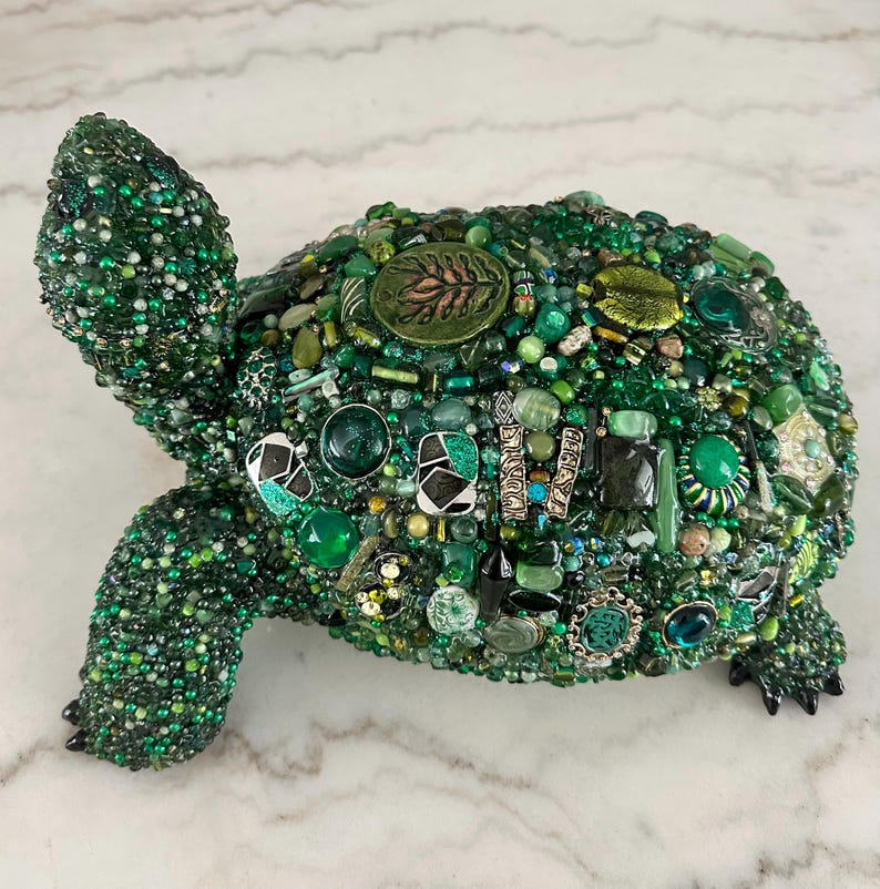 May include: A decorative turtle sculpture covered in various shades of green beads, gems, and decorative elements. The turtle's shell is adorned with a variety of green stones and embellishments. The sculpture has a detailed head, legs, and tail.