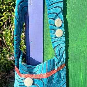 Może przedstawiać: Turkusowa torebka z aksamitu z czarnymi falistymi liniami i czerwoną wstążką. Torebka ma trzy białe guziki.