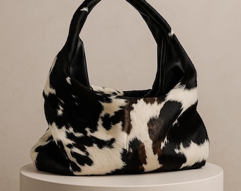 May include: A hobo-style handbag featuring a black and white cowhide pattern with a soft, textured fur finish. The bag has a single integrated handle, a slouchy, unstructured silhouette, and a smooth black interior lining visible at the opening.