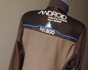 Puede incluir: Una chaqueta negra con una tira de luz azul alrededor del cuello y una tira de luz azul alrededor de la parte inferior de la chaqueta. La chaqueta tiene un parche blanco en la espalda que dice "ANDROID Designed by CYBERLIFE Made in DETROIT" y un triángulo blanco con una luz azul en su interior. Debajo del triángulo está el texto "RK800". La chaqueta tiene una tira de luz azul en la manga derecha.