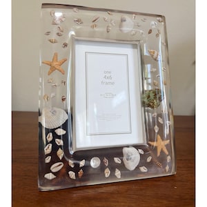 Puede incluir: Un marco de fotos rectangular transparente con conchas marinas y estrellas de mar incrustadas en el borde. El inserto central muestra el texto one 4x6 frame y genuine SONOMA home decor. El marco está diseñado para una foto de 10x15 cm.