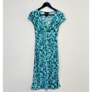 May include: A knee-length dress with a V-neck and short sleeves, patterned with blue, green, and purple abstract shapes. The dress hangs on a black hanger against a white backdrop. The dress has a fitted bodice and a slightly flared skirt.
