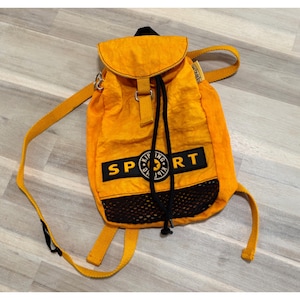May include: Bright yellow backpack with black accents and the word "SPORT" in black letters. The backpack has a drawstring closure, a flap, and a mesh pocket. The backpack has a shoulder strap and two smaller straps.