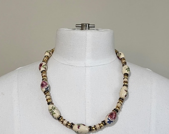 May include: Cream-colored beaded necklace with floral designs. The necklace features oval-shaped beads with pink and red roses, interspersed with gold and cream-colored beads. The necklace is displayed on a white mannequin.