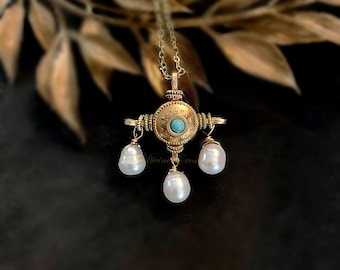 May include: A gold-toned pendant necklace with a central turquoise stone and three dangling white pearls. The pendant has an ornate, circular design with intricate detailing. The necklace hangs on a delicate chain.