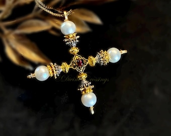 May include: Gold-toned cross pendant necklace with four white pearl accents and a central red gemstone. The cross features ornate detailing and is suspended from a delicate gold chain. The background is dark, highlighting the jewelry.