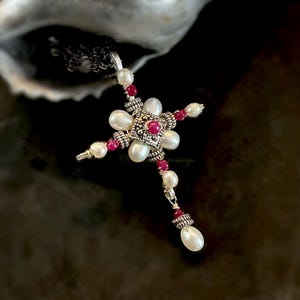 May include: A cross pendant featuring white oval pearls and small round red gemstones set in silver-toned metal. The center square component has an intricate textured pattern with a central red stone, surrounded by pearls and red accents on a chain.