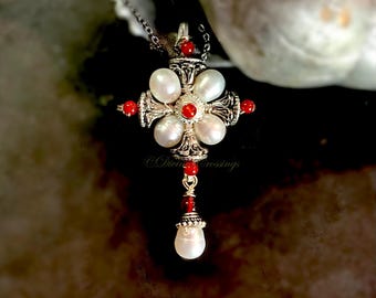 May include: A cross-shaped pendant featuring four white oval pearls arranged around a central red bead, accented with smaller red beads and intricate silver-toned metalwork. A single teardrop pearl hangs from the bottom on a silver chain.