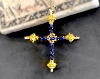 May include: A gold-toned cross pendant featuring deep blue faceted stones embedded along the arms. The center and ends of the cross are adorned with ornate gold-toned metal caps. The piece hangs from a delicate gold-toned chain and includes the text Divine Crossings.