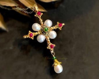 May include: A gold-toned cross pendant necklace featuring four white oval pearls arranged in a flower pattern around a central green stone. The cross is accented with small red stones at each point and a single dangling white pearl at the bottom.