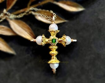 May include: Gold-colored cross pendant necklace with white pearls at the ends of the cross and a green gem in the center. The necklace is on a gold chain. The background is dark with gold-colored leaves.