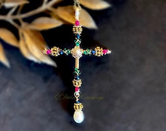 May include: A gold-toned cross pendant necklace with red, green, and blue crystal accents. The cross features a pearl at the center and a dangling pearl at the bottom. The text "Divine Crossings" is visible.