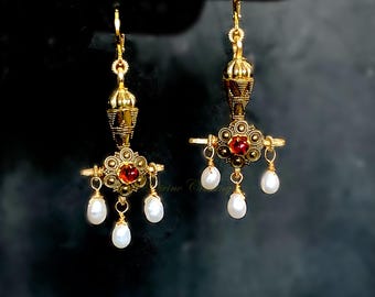 May include: A pair of gold-toned drop earrings. Each earring features a central red gemstone surrounded by smaller gold circles, with three dangling white pearls. The earrings have an ornate, vintage design.
