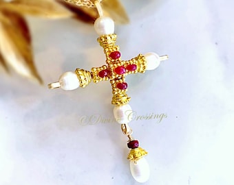 May include: A gold cross pendant necklace featuring a textured metal frame, four white pearls at the ends of the cross arms, and small red gemstones embedded in the center. A dangling pearl and red gemstone accent hangs from the bottom of the cross.