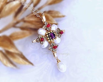 May include: A gold-toned cross pendant necklace featuring a central purple gemstone, four white oval pearls, and four small red stones set within ornate leaf-shaped metalwork. A single white teardrop pearl hangs from the base of the cross.