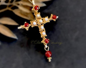May include: A gold-toned cross pendant featuring a textured, beaded metal surface. The cross is embellished with small, round white pearls and faceted red stones at the ends and center. A single red bead hangs from the bottom of the cross.