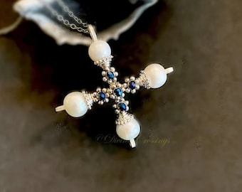 May include: A silver-toned cross pendant necklace with four large, round, white pearls at the ends and smaller blue gemstones at the center. The necklace has a delicate chain and is set against a dark background.