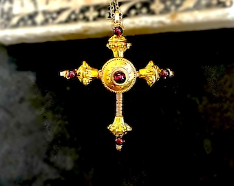 May include: A gold-colored pendant necklace featuring a cross design. The cross has a central circular element and four arms, each adorned with a red gemstone. The pendant hangs from a delicate chain, showcasing intricate details and a vintage aesthetic.