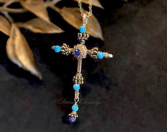 May include: Gold-toned cross pendant necklace with turquoise and blue gemstone accents. The cross features intricate wirework and a central blue gemstone. The necklace is suspended from a delicate gold chain. The text "Divine Crossings" is visible.