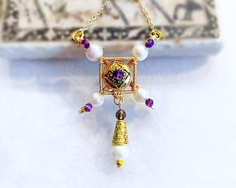 May include: A gold-toned necklace featuring a square wire-wrapped pendant with a central purple gemstone, surrounded by white pearls and smaller purple beads. A dangling gold-toned cone charm with a pearl and dark bead hangs from the bottom center.
