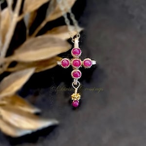 May include: A silver-toned cross pendant necklace with ruby-colored gemstones. The cross features five round gems on the cross arms and three vertically aligned gems. A small gold-toned bead cluster and a single ruby dangle from the bottom. The text "Divine Crossings" is visible.