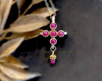 May include: A silver-toned cross pendant necklace with ruby-colored gemstones. The cross features five round gems on the cross arms and three vertically aligned gems. A small gold-toned bead cluster and a single ruby dangle from the bottom. The text "Divine Crossings" is visible.