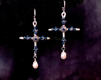 May include: Silver-toned cross earrings with blue crystal accents and a dangling pearl. The cross design features intricate wire wrapping and a central blue stone. The earrings have a hook closure.