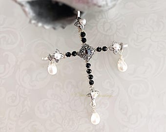 May include: A silver-toned cross pendant featuring black beads along the arms, a central dark square stone, and three white teardrop-shaped pearls dangling from the ends. The metalwork has an intricate, textured finish with small floral accents.