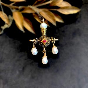 May include: A gold-toned metal cross pendant featuring an intricate filigree design with a central red stone. The pendant is adorned with four white teardrop-shaped pearls, one at the top and three hanging from the base, suspended on a thin chain.