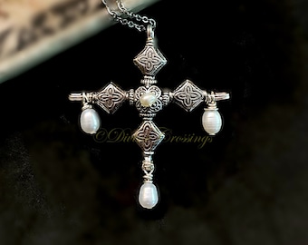 May include: A silver-toned metal cross pendant on a delicate chain, featuring intricate embossed floral patterns on each arm. The center and three ends of the cross are adorned with small, white, oval-shaped freshwater pearls hanging from wire.