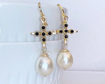 May include: A pair of gold-tone drop earrings featuring a cross-shaped charm adorned with small black stones. Each cross is suspended from a gold-tone hook and hangs above a single, large, teardrop-shaped white pearl with a decorative gold cap.