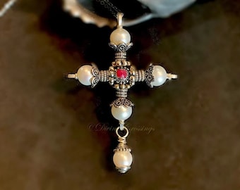 May include: A silver-toned cross pendant necklace with a central red gemstone and four white pearls. The cross has ornate detailing and is suspended from a delicate chain. The pendant is a religious symbol.