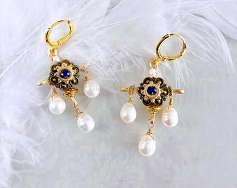 May include: A pair of gold-colored earrings with a floral design and a dark blue center stone. Each earring features three dangling white pearls and a lever back closure. The earrings are set against a white background with feathers.