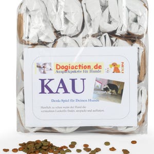 May include: A clear plastic-wrapped package containing dog treats and a white card with the text "KAU" and "Dogiaction.de". The package includes brown and white paper packaging. A small pile of dog food is in front of the package.