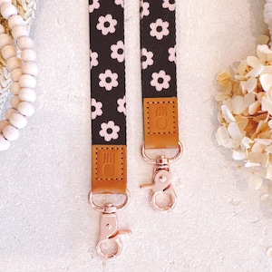 May include: Two black and white floral patterned straps with rose gold clasps. The straps have brown leather accents with the initials "NM" embossed. The background is a soft white with decorative elements.