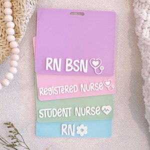 Peut inclure: Une pile de quatre porte-badges rectangulaires en plastique aux couleurs pastel. La carte violette du haut porte l'inscription RN BSN avec une icône de stéthoscope. La carte rose indique REGISTERED NURSE avec un stéthoscope. La carte verte indique STUDENT NURSE avec un cœur. La carte bleue indique RN avec une icône de fleur.
